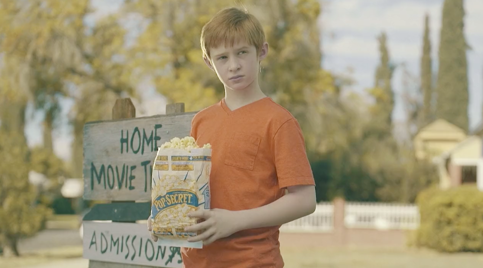 Boy in an orange shirt holding a bright yellow Pop Secret popcorn bag